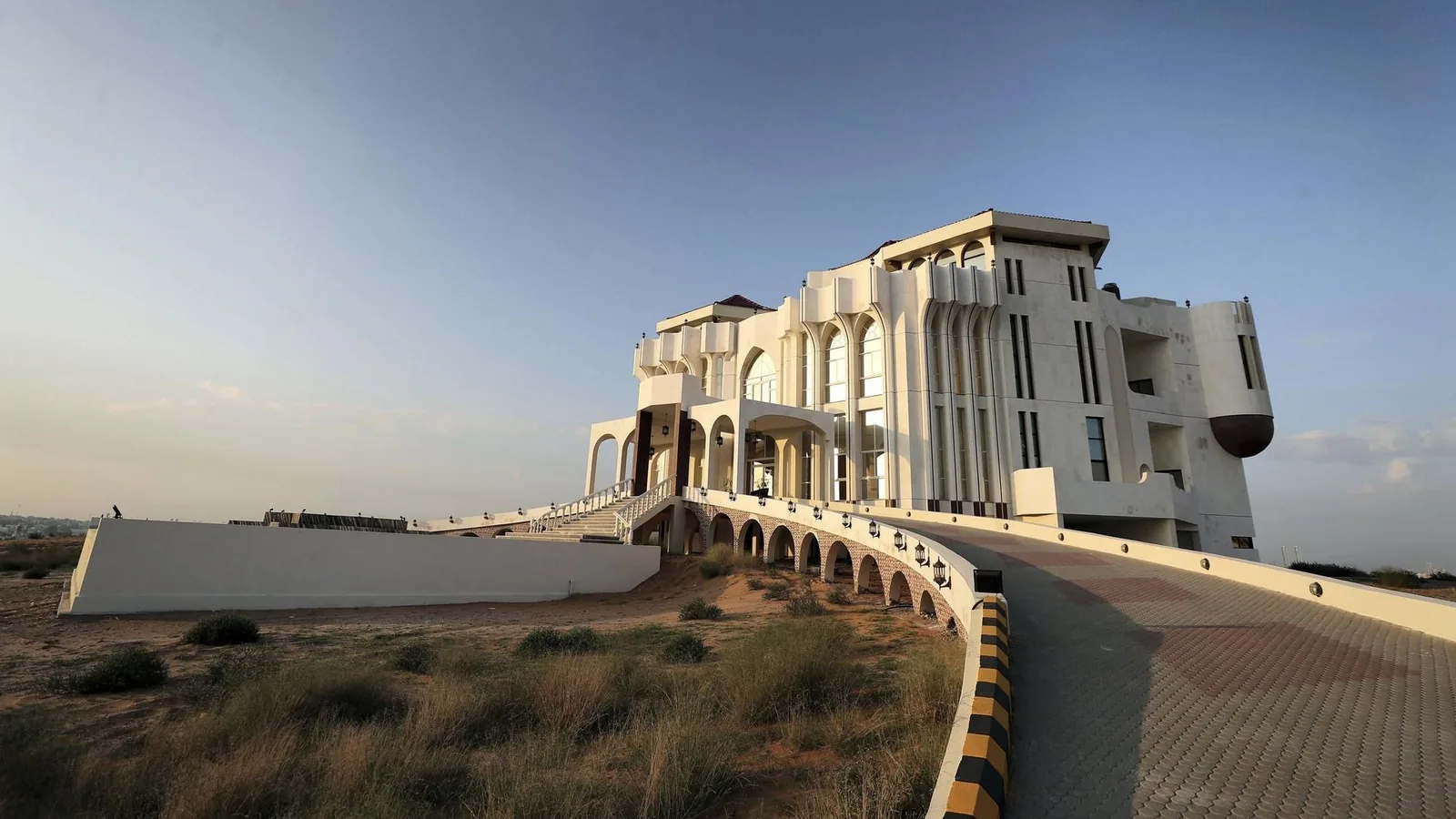 A grand white palace-style building with pointed arched windows, arched arcades, and grand staircases, set on a low hill above sandy desert with a curving tiled driveway leading up to it under a clear sky.