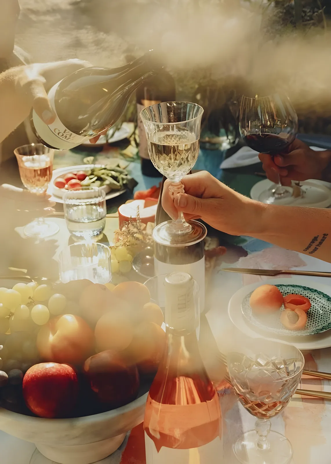 A hand holds a crystal wine glass being filled with white wine while a bowl of grapes and peaches, a bottle of rosé, and plates of food sit on a table in warm hazy sunlight.