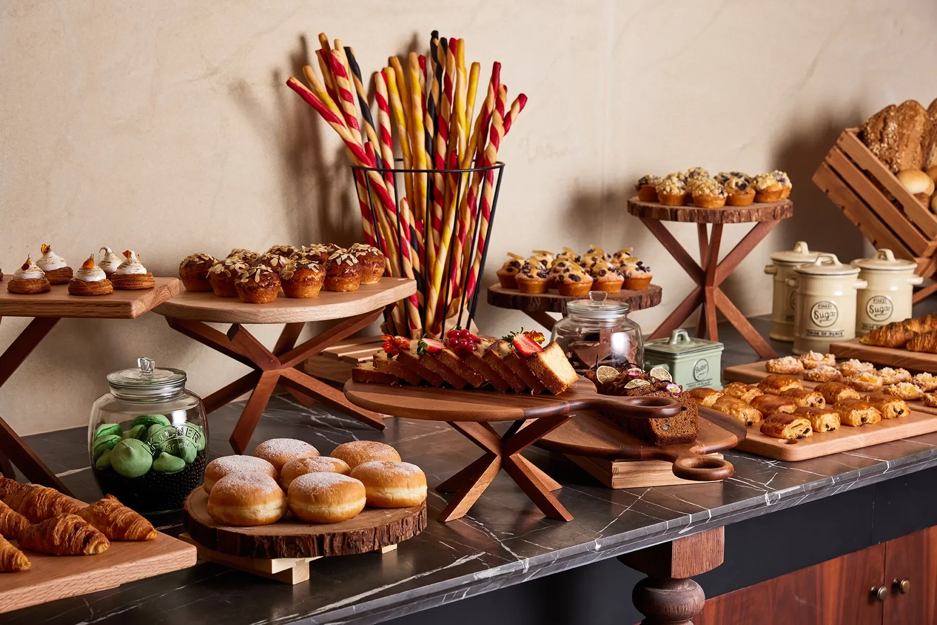 A hotel buffet on a black marble counter with wooden tiered stands holding almond muffins, croissants, pain au chocolat, sugar-dusted doughnuts, sliced pound cake topped with strawberries and berries, green macarons in a glass jar, striped bread sticks in a wire basket, ceramic sugar canisters, and a crate of loaves.