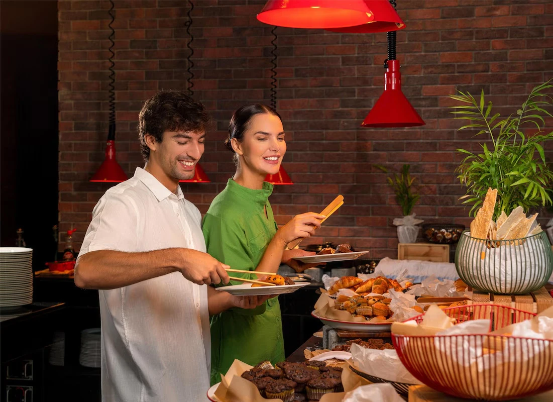Radisson Resort Ras Al Khaimah Marjan Island, Al Hamra, Ras Al Khaimah: A man in a white shirt and a woman in a green dress hold plates and use serving tongs at a buffet spread of muffins, croissants, and pastries, with red heat lamps and a brick