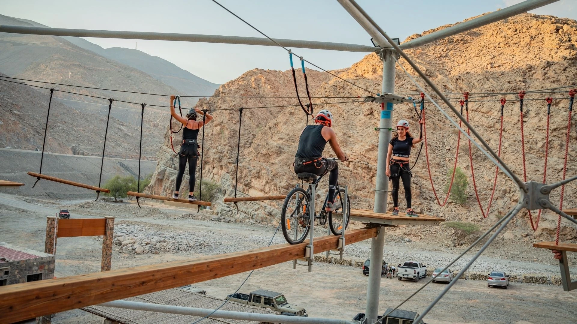 Jais Ropes Course Ras Al Khaimah, Ras Al Khaimah: A man rides a bicycle across a wooden beam on an elevated high ropes course while two women in helmets and harnesses balance on nearby planks, with rocky brown mountains and a parking area in the