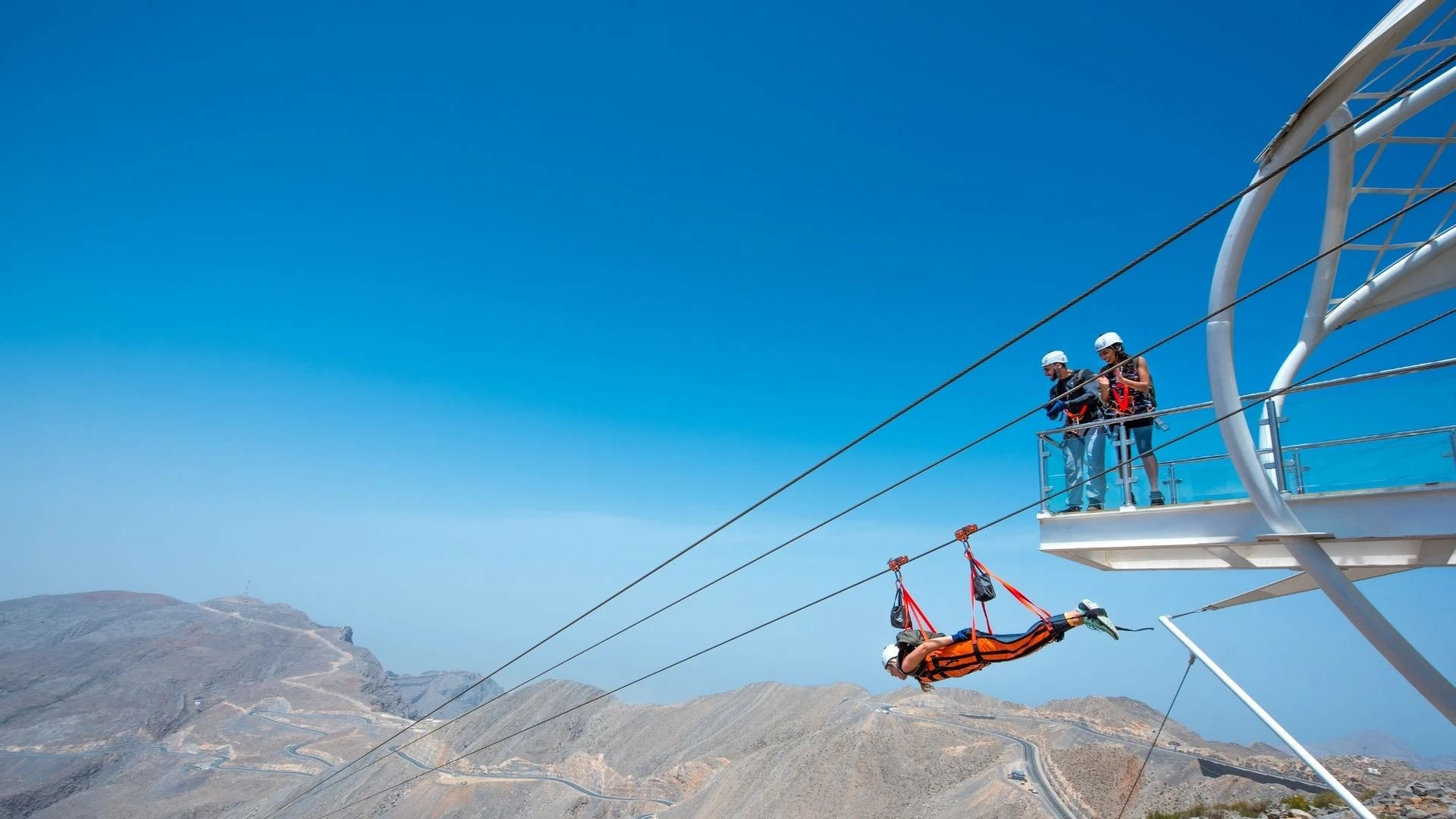 A person in an orange harness ziplines face-down along steel cables above brown rocky mountains, with two helmeted people and a staff member on a metal launch platform on the right beneath a bright blue sky.
