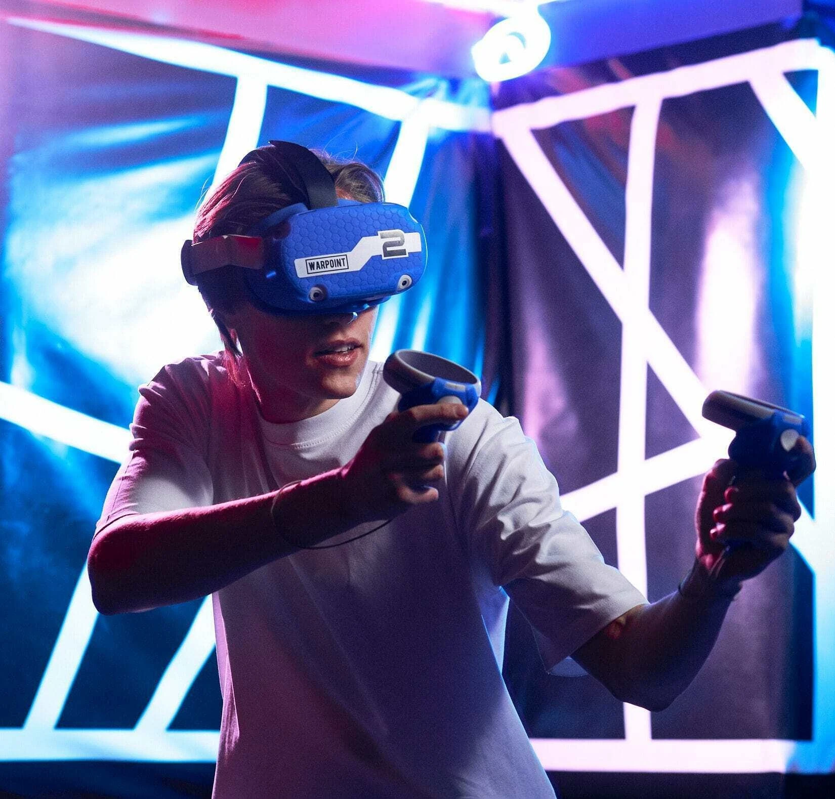 Too Hot Outside? Try These 7 Indoor Activities in Ras Al Khaimah this Long Weekend, Ras Al Khaimah: A person wearing a blue VR headset labeled Warpoint holds two black motion controllers, standing in a dark room lit by blue and pink neon light