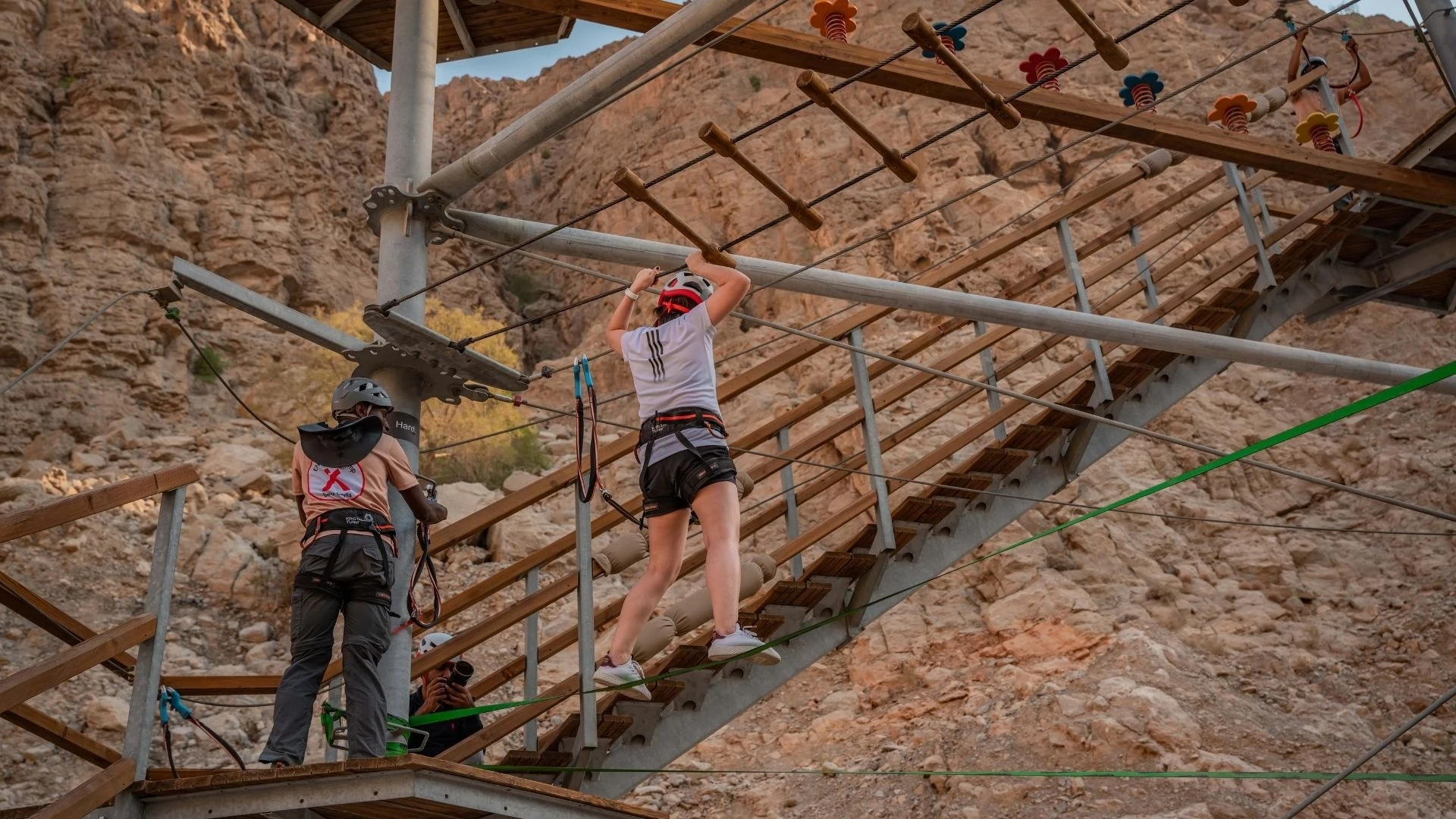 Jais Ropes Course Ras Al Khaimah, Ras Al Khaimah: A person wearing a red helmet, white shirt, black shorts, and a climbing harness crosses a suspended wooden and cable obstacle on an outdoor high ropes course, with a staff member in a peach shirt