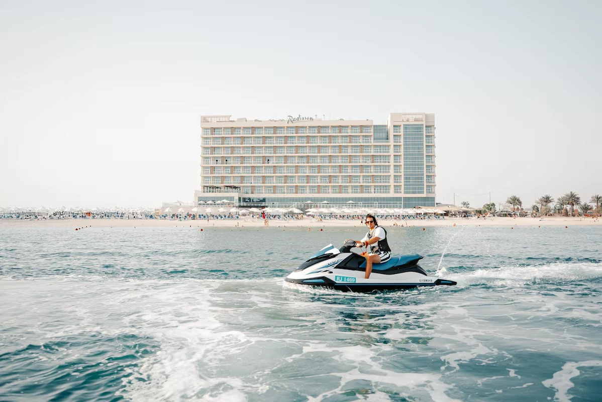 Radisson Resort Ras Al Khaimah Marjan Island, Al Hamra, Ras Al Khaimah: A rider on a blue and white Yamaha jet ski marked RJ 1469 speeds across turquoise sea, with a wide beachfront hotel, umbrellas, and sunbeds along the sandy shore behind.