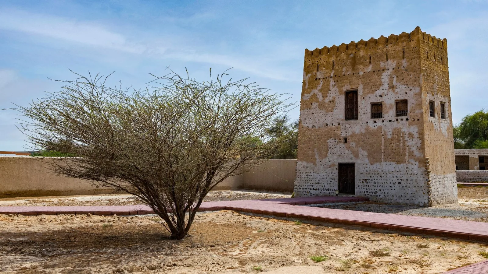 A square historic watchtower built of stone and mud with small wooden windows and a crenellated roofline, set in a sandy enclosure with a red brick path and a leafless spreading acacia tree in the foreground under a partly cloudy sky.