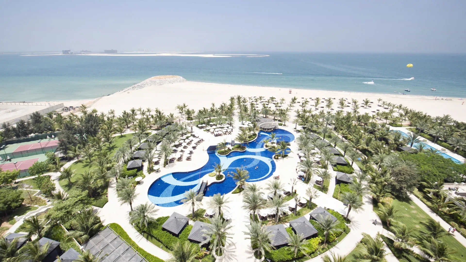 Waldorf Astoria Ras Al Khaimah, Al Hamra, Ras Al Khaimah: Aerial view of a beach resort featuring a curving blue mosaic swimming pool surrounded by palm trees, thatched cabanas, and manicured lawns, with a wide sandy beach and calm turquoise sea and
