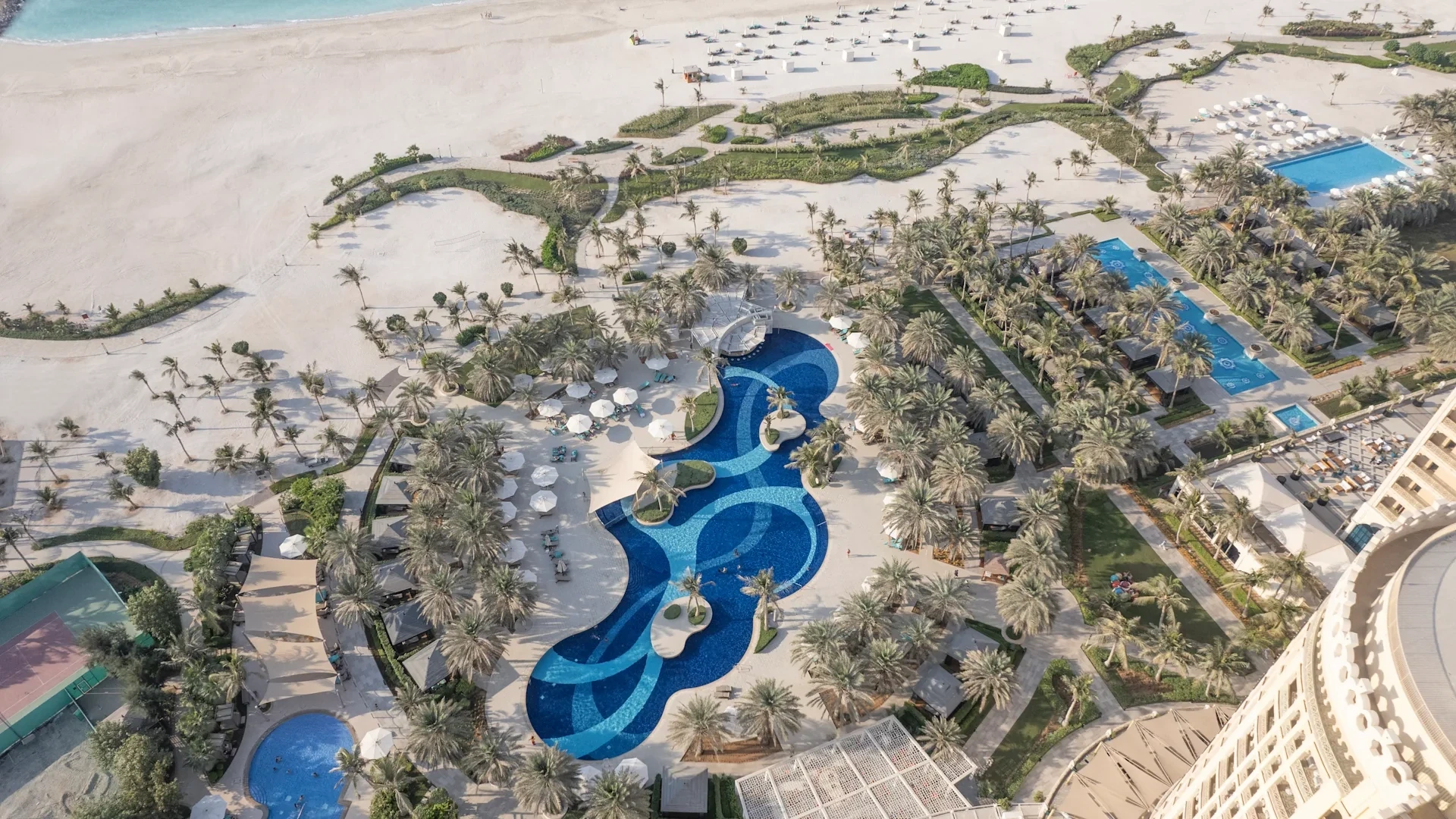 Waldorf Astoria Ras Al Khaimah, Al Hamra, Ras Al Khaimah: Aerial view of a beach resort featuring a large curving blue swimming pool with mosaic patterns, clusters of palm trees, white umbrellas, additional pools, and a wide sandy beach beside the