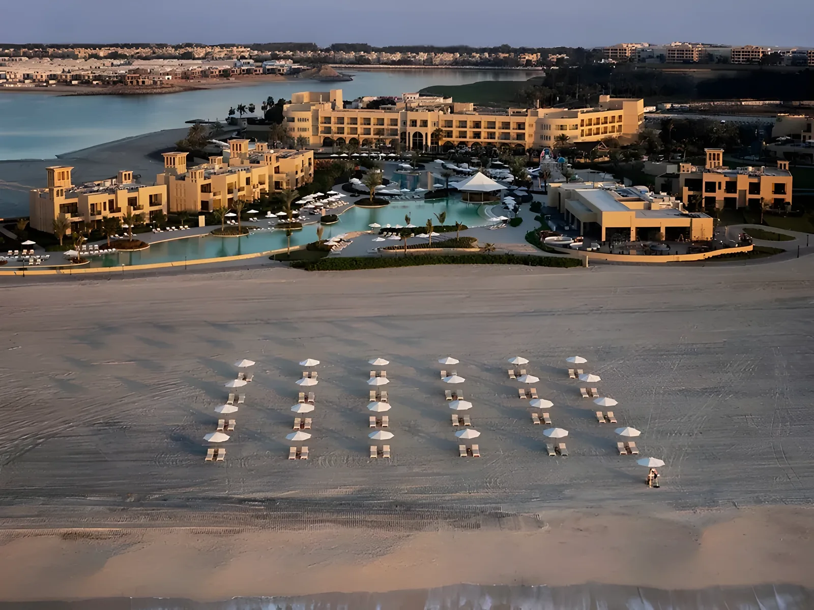 Aerial view of a beachfront resort featuring rows of white parasols and sun loungers arranged on a wide sandy beach, with a lagoon-style swimming pool and tan low-rise buildings beside a calm bay in warm evening light.