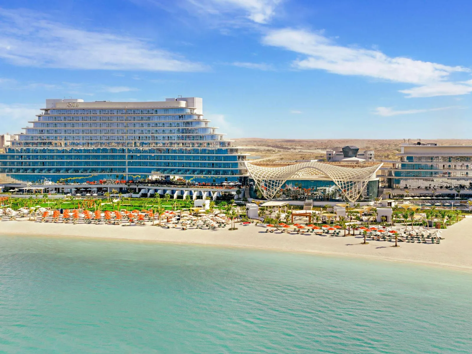 Aerial view of a tiered glass-fronted beachfront resort with a curved white pavilion, rows of orange and white sun umbrellas on white sand, turquoise sea in the foreground, and desert hills in the background under a clear blue sky.