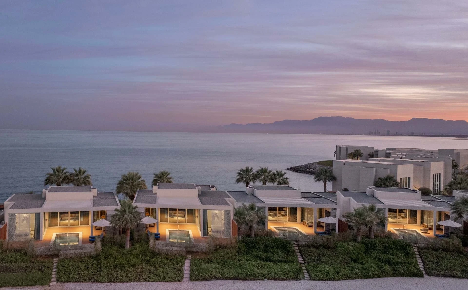 InterContinental Ras Al Khaimah Mina Al Arab Resort & Spa, an IHG Hotel, Mina Al Arab, Ras Al Khaimah: Aerial view of white single-story beachfront villas with lit private pools and palm trees, facing a calm sea at sunset with mountains and a pastel