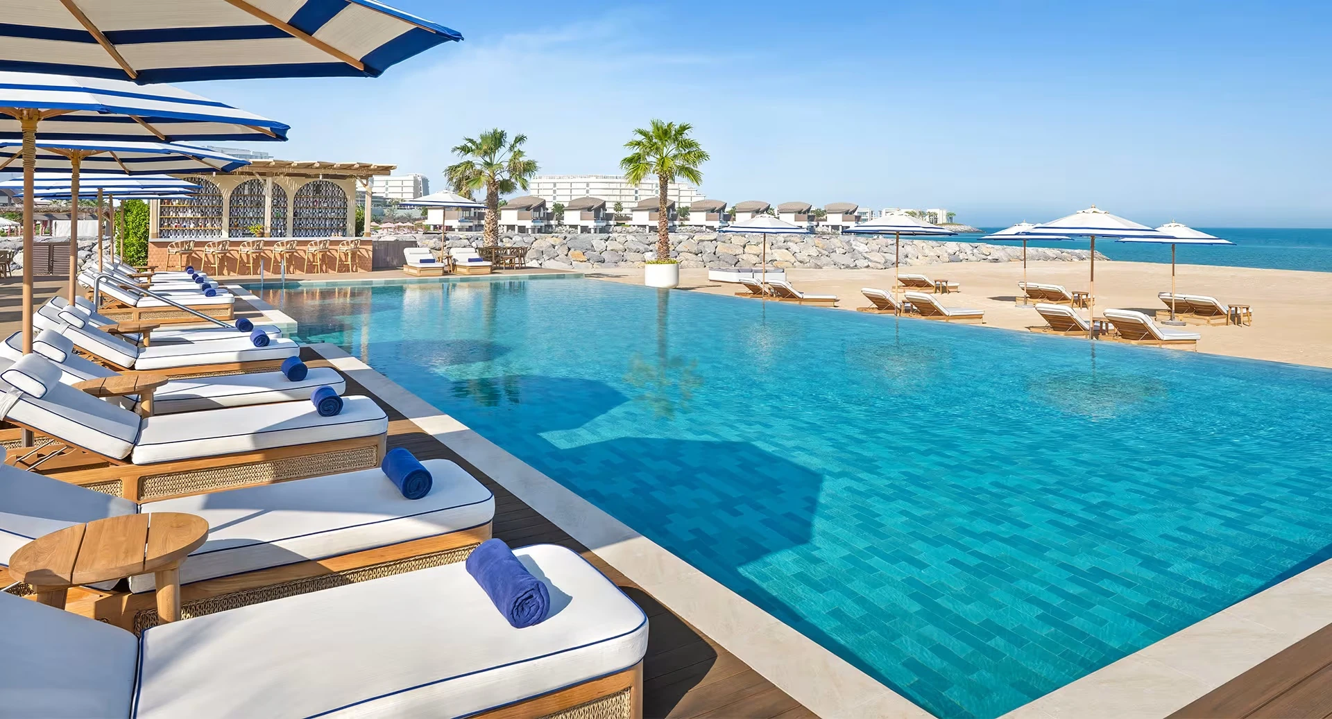 Anantara Mina Ras Al Khaimah Resort, Mina Al Arab, Ras Al Khaimah: Beachfront infinity pool with turquoise water, a row of white cushioned sun loungers with rolled blue towels, blue-and-white striped umbrellas, palm trees, a poolside bar, and the