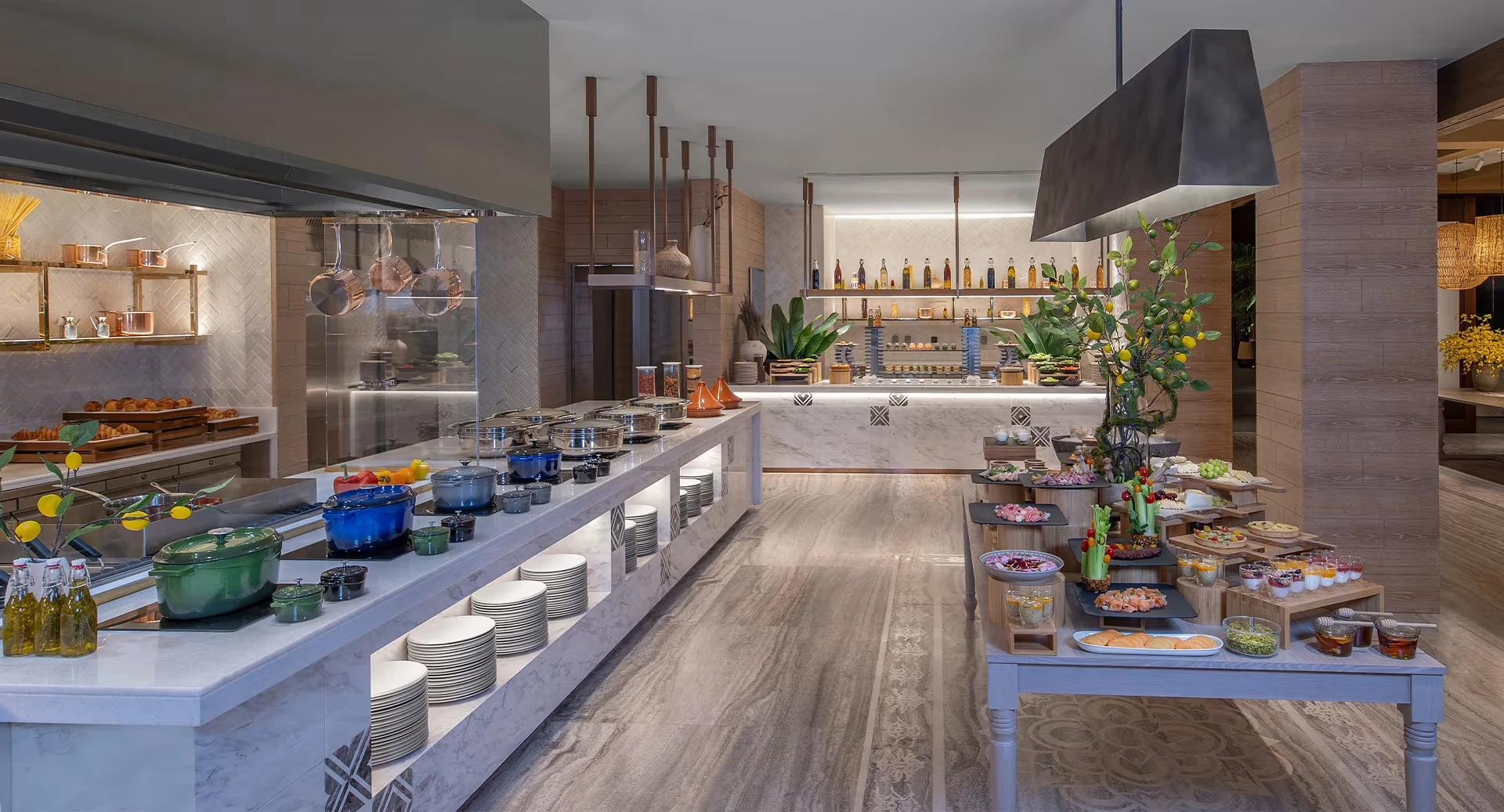 Anantara Mina Ras Al Khaimah Resort, Mina Al Arab, Ras Al Khaimah: Buffet restaurant with a long marble counter holding stacked white plates, blue and green cast iron pots, and chafing dishes on the left, while a wooden table on the right displays