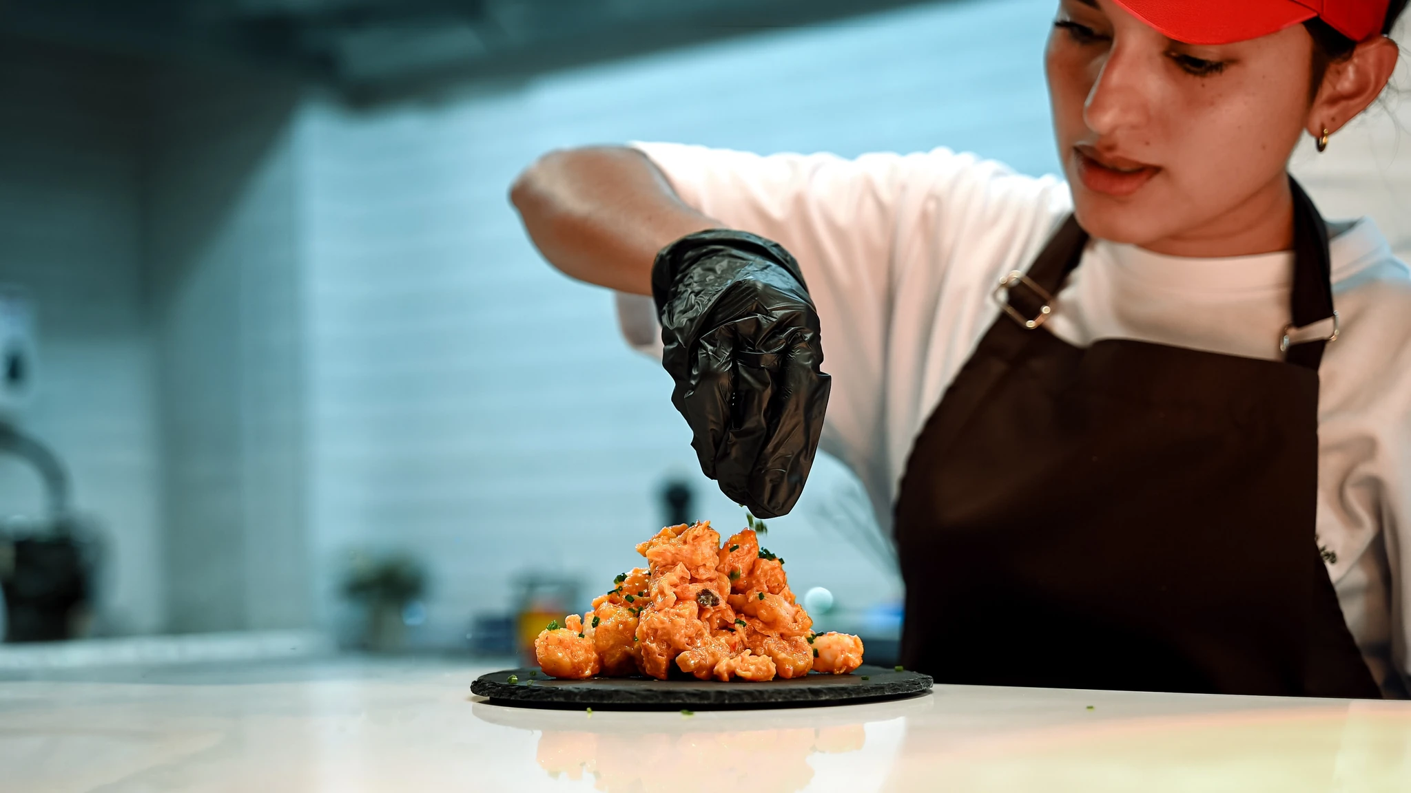 Chef wearing a black glove, brown apron, and red cap sprinkling green herbs over a mound of saucy shrimp arranged on a round black slate plate atop a white counter.