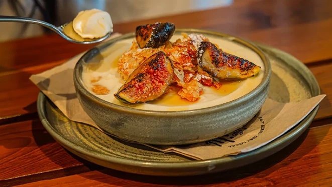 Close-up of a grey ceramic bowl holding a pale creamy dessert topped with roasted fig quarters, honeycomb pieces, and drizzled honey, set on a matching plate over brown paper on a wooden table, with a spoon of cream held above.