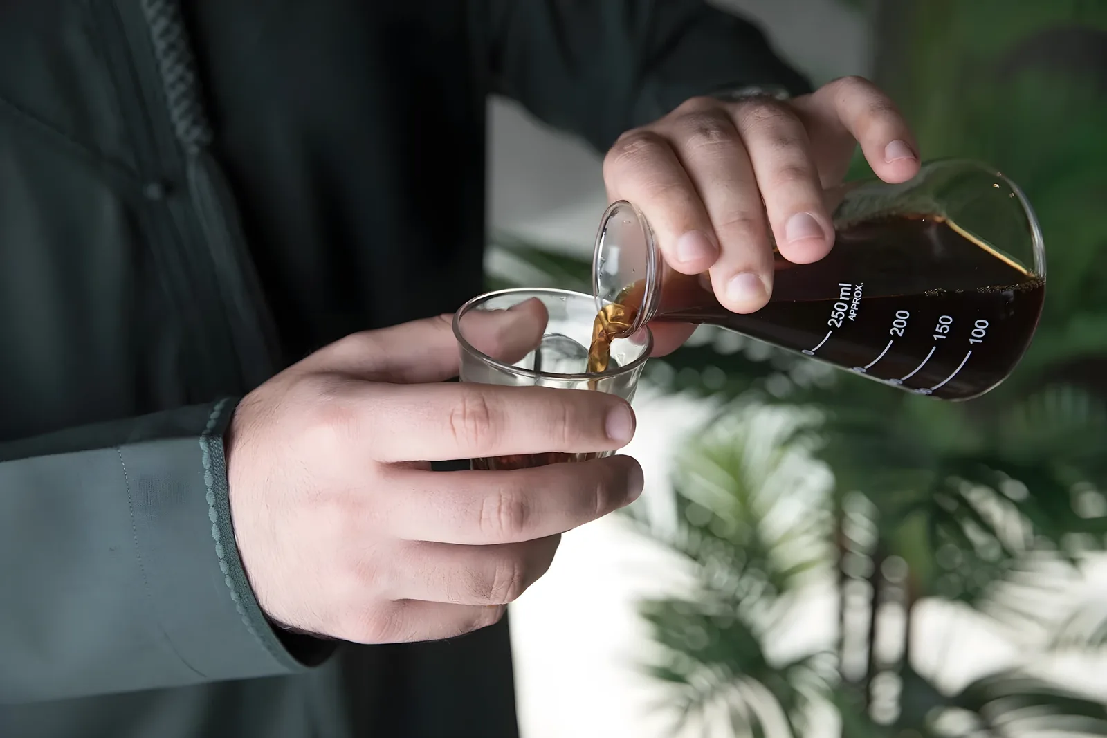 Close-up of a man's hands pouring dark brown coffee from a 250ml graduated glass beaker into a small clear glass cup, tropical green plants blurred behind.