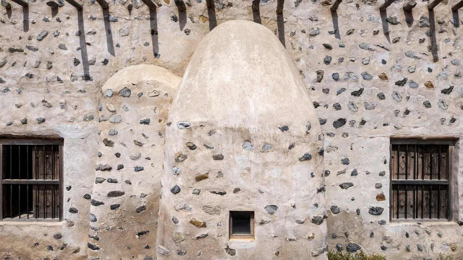 Close-up of a pale stone and lime plaster wall embedded with grey rounded stones, featuring a domed egg-shaped structure, a small square window, and two dark wooden barred windows, with projecting wooden roof beams along the top.
