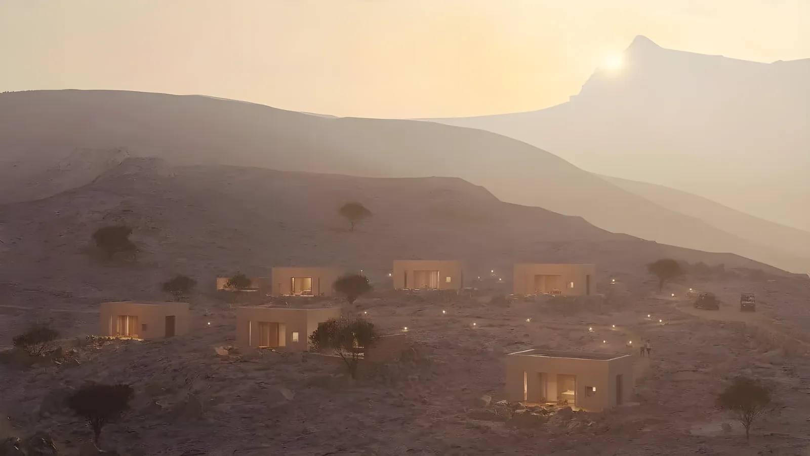 Cluster of flat-roofed sandstone villas with lit windows scattered across a rugged desert mountainside, sparse acacia trees, and a hazy sunset over layered peaks in the background.