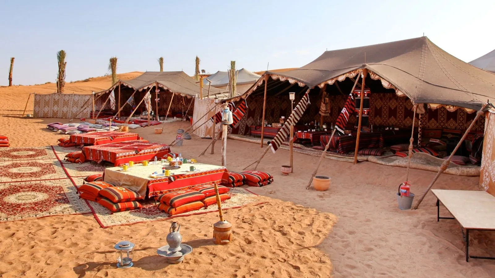Desert camp featuring canvas Bedouin tents, red and orange floor cushions, low tables with cloth coverings, patterned carpets, and metal coffee pots on the sand under a clear sky with dunes behind.