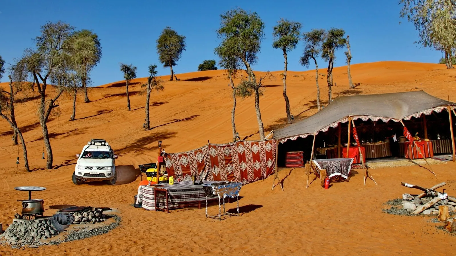 Desert camp with a large open Bedouin tent, red and white patterned windbreak fabrics, a low seating table, a white four-wheel-drive vehicle, ghaf trees, orange sand dunes, and a stone fire pit under a clear blue sky.