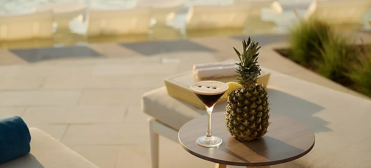 Espresso martini in a stemmed glass with a pineapple wedge garnish, next to a whole pineapple on a round wooden table, with sun loungers and blurred pool water in the background.