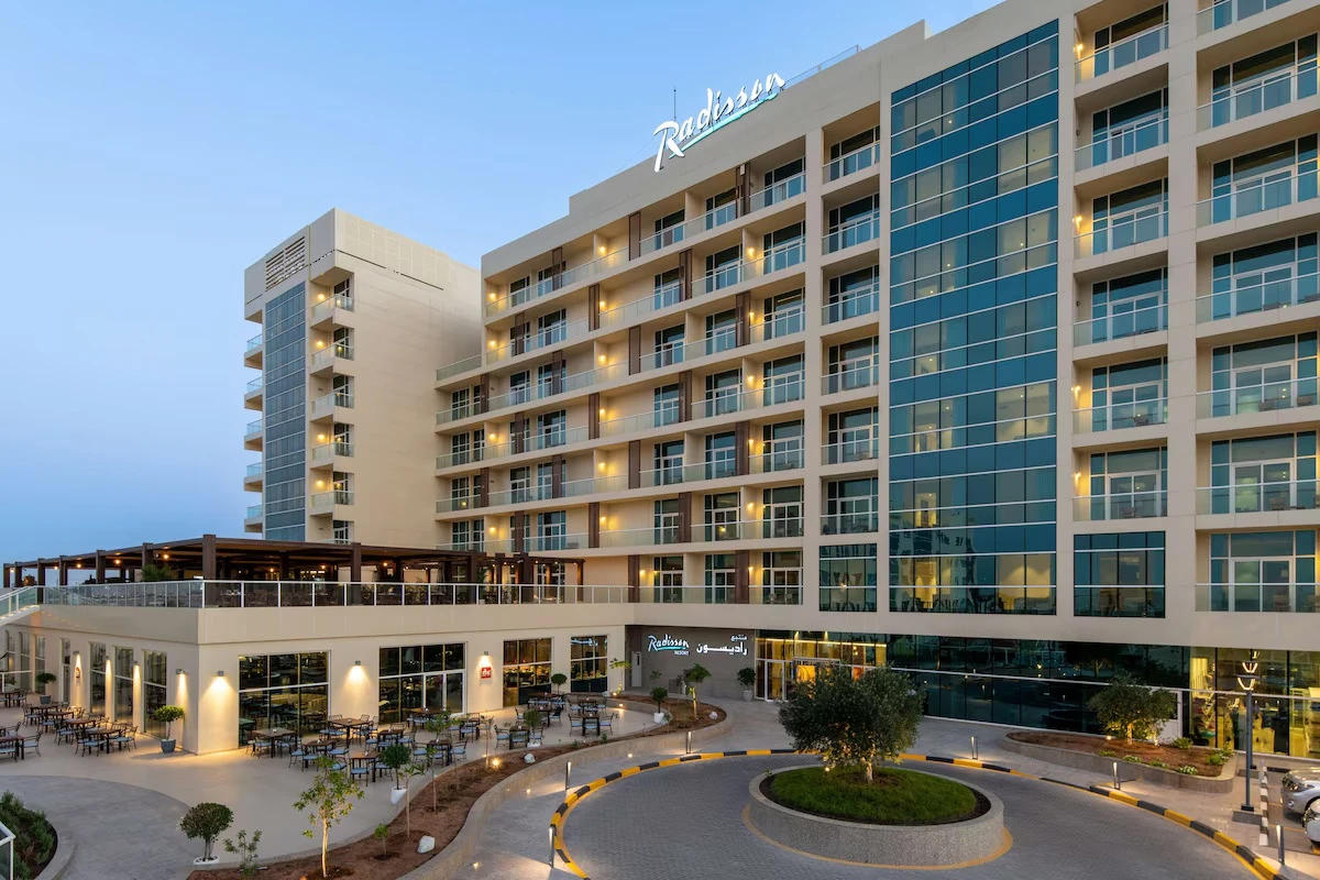 Radisson Resort Ras Al Khaimah Marjan Island, Al Hamra, Ras Al Khaimah: Exterior of a Radisson resort hotel at dusk showing a tall cream and blue glass building with balconies, an illuminated Radisson sign, a curved paved driveway with landscaped
