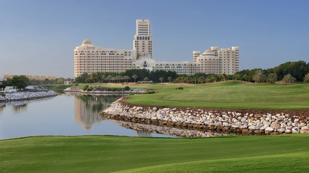 Waldorf Astoria Ras Al Khaimah, Al Hamra, Ras Al Khaimah: Green golf fairway beside a still lake with a stone bank, framing a large domed cream-colored resort and tower in the background under a clear sky.