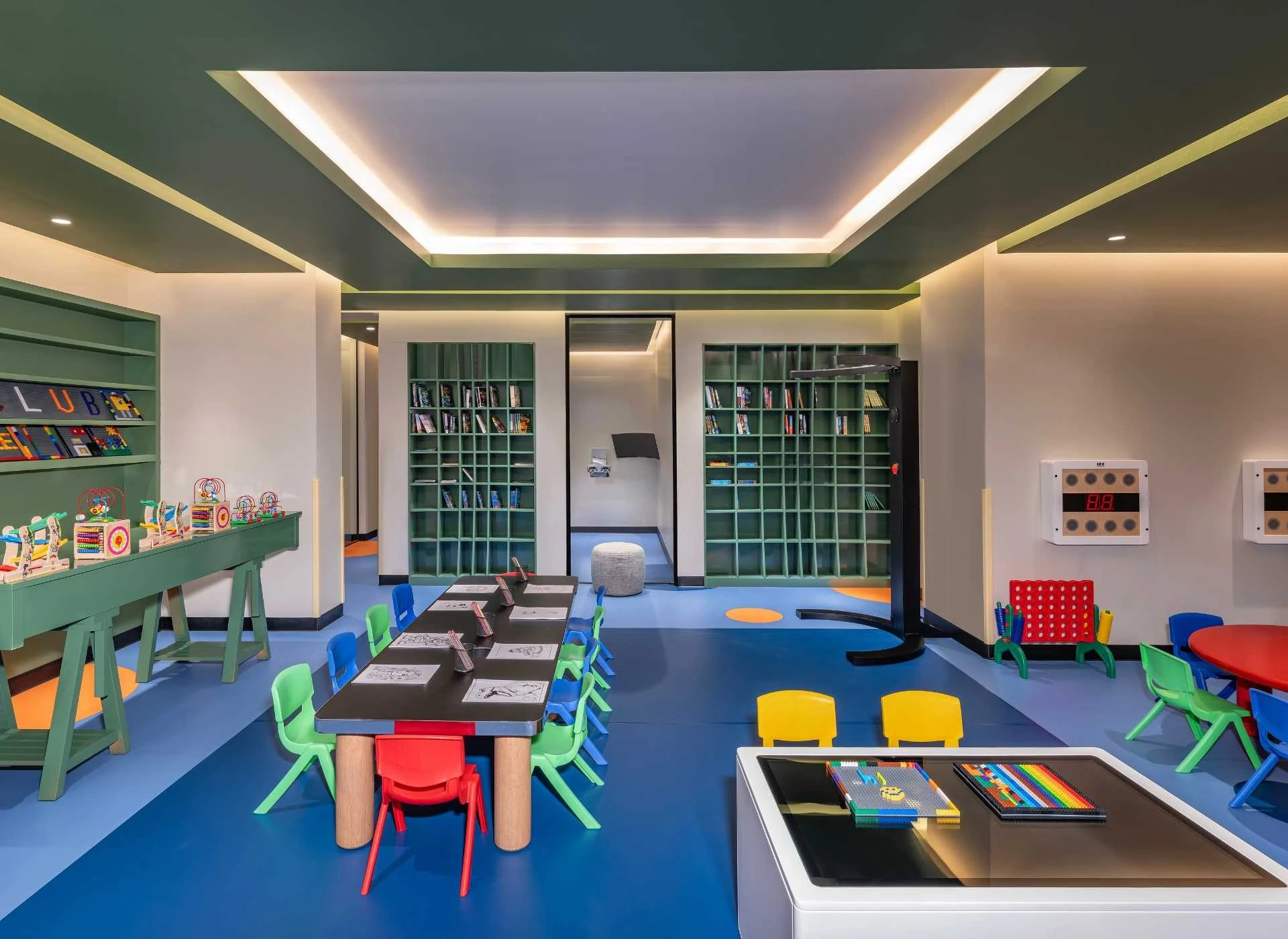 InterContinental Ras Al Khaimah Mina Al Arab Resort & Spa, an IHG Hotel, Mina Al Arab, Ras Al Khaimah: Indoor children's play area with a long black table surrounded by red, green, blue, and yellow plastic chairs, green shelving units filled with
