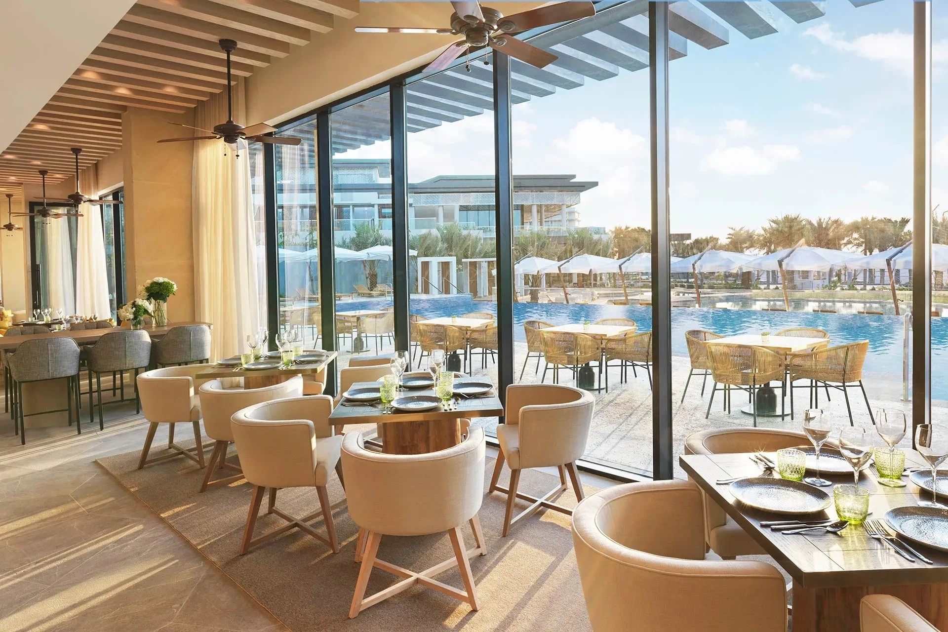 InterContinental Ras Al Khaimah Mina Al Arab Resort & Spa, an IHG Hotel, Mina Al Arab, Ras Al Khaimah: Interior of a resort restaurant with cream upholstered chairs, wooden tables set with plates and green glassware, ceiling fans, and full-height