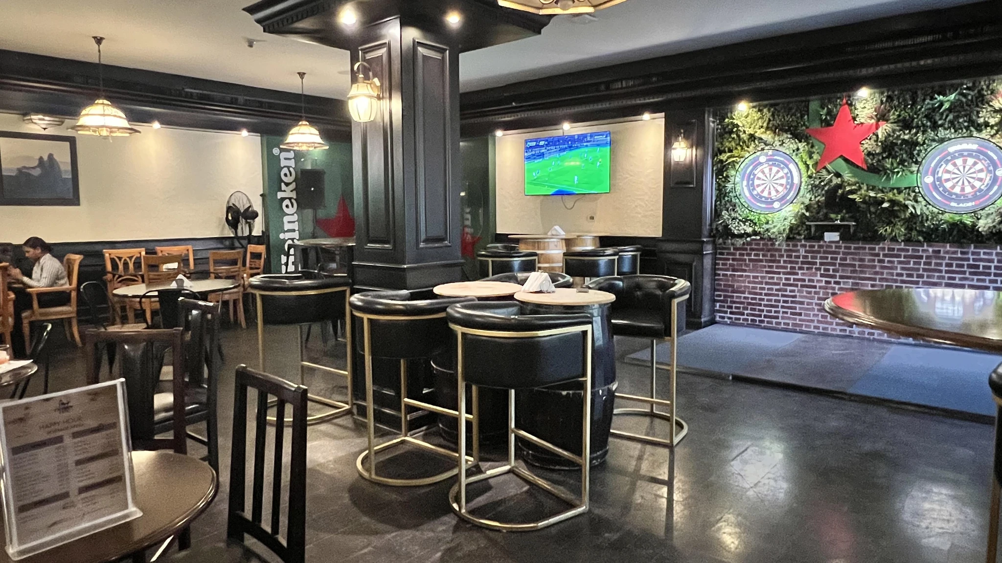 Interior of a sports bar with black leather tub chairs at barrel tables, a large screen showing a football game, two dartboards on a green plant wall, brick accents, pendant lights, and wooden dining chairs on the left.