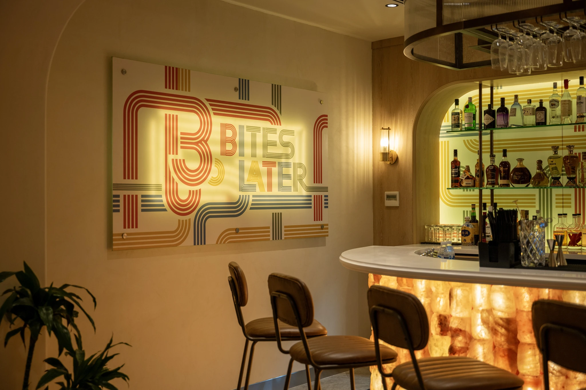 Interior of a stylish bar with a retro striped Bites Later sign in red, blue, and yellow on a beige wall, an arched shelf holding liquor bottles, hanging glassware, a backlit onyx bar counter, and brown leather bar stools.