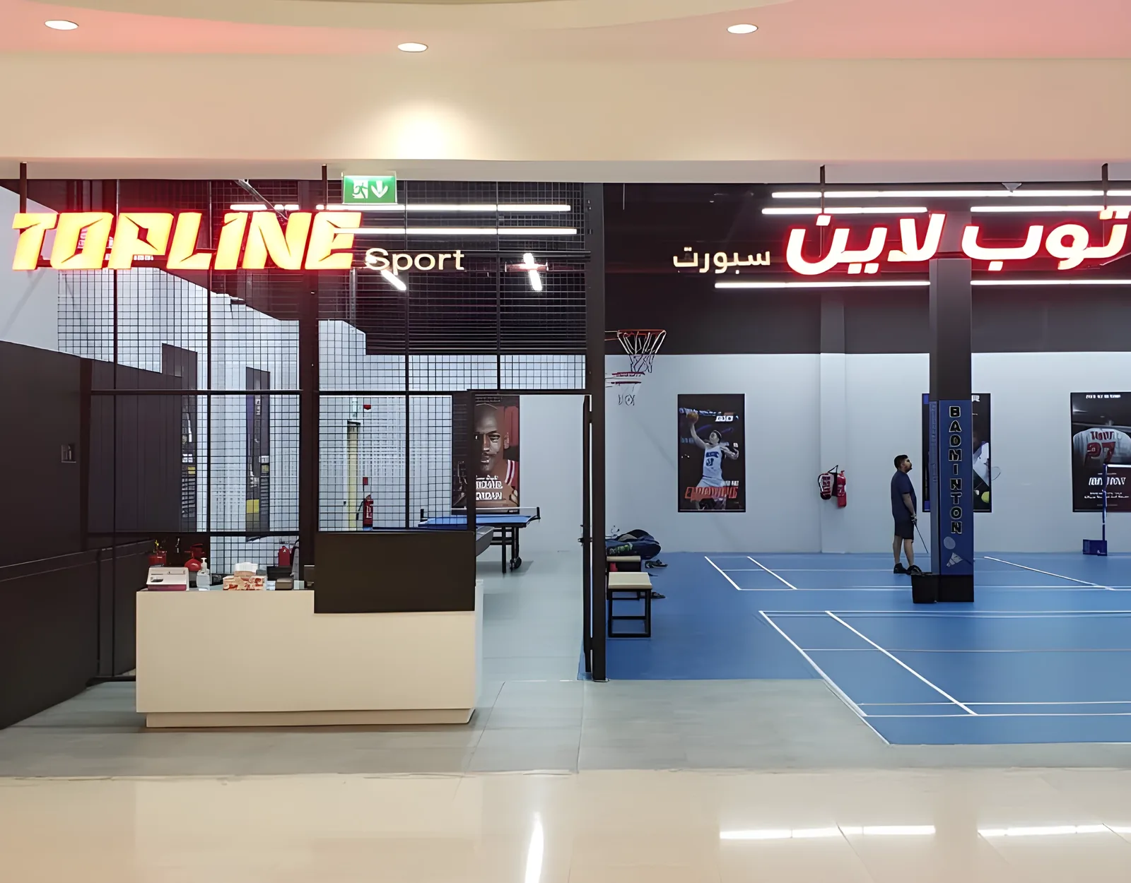 Interior of an indoor sports facility with a red neon sign reading Topline Sport in English and Arabic, a blue badminton court with a person standing on it, a basketball hoop, table tennis table, and a reception counter in the foreground.