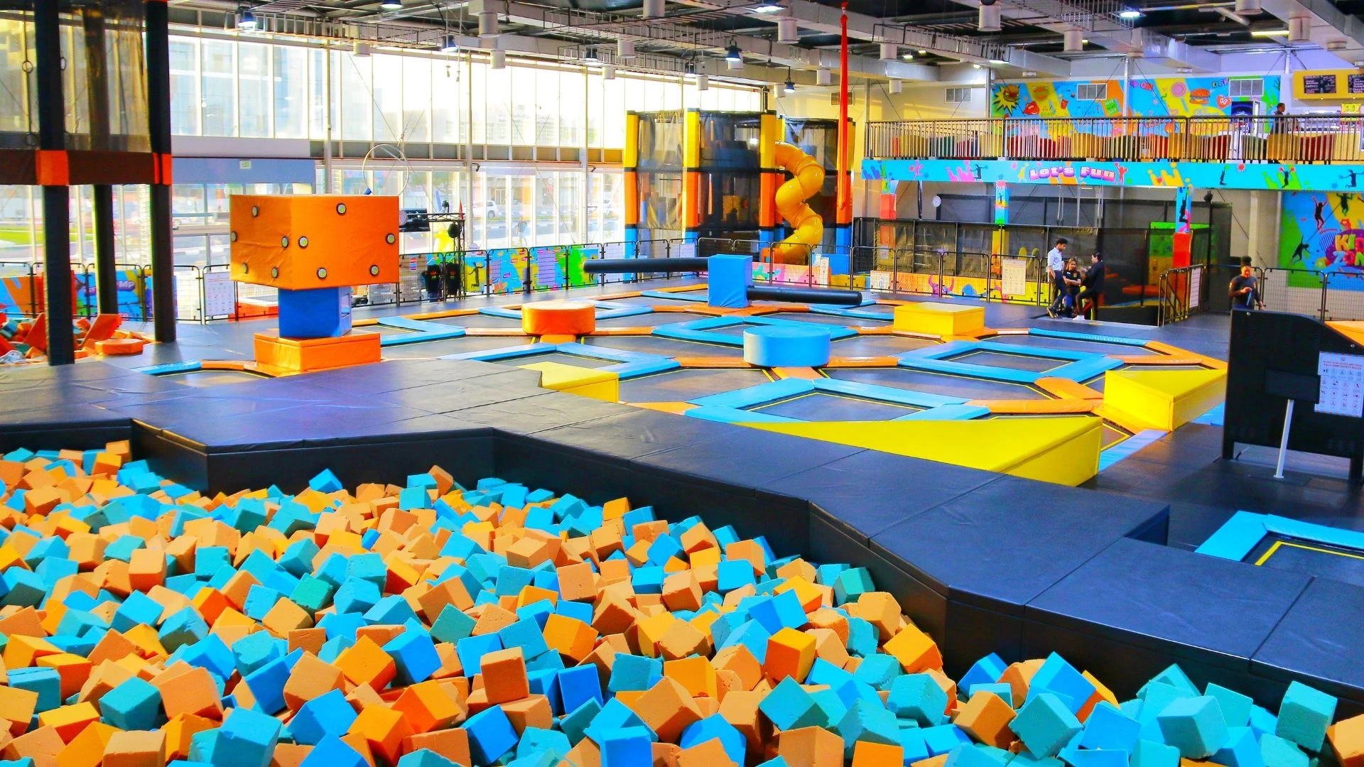 Too Hot Outside? Try These 7 Indoor Activities in Ras Al Khaimah this Long Weekend, Ras Al Khaimah: Interior of an indoor trampoline park with a foam pit full of blue and orange foam cubes in the foreground, rows of trampolines, padded obstacles, a