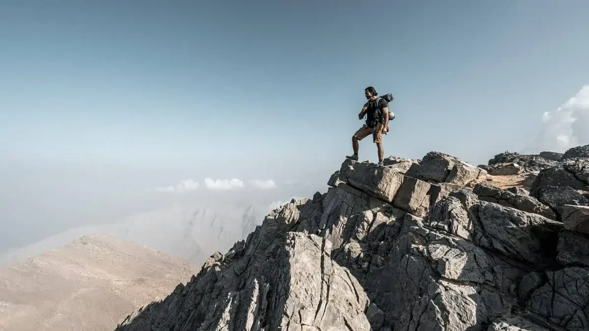 A man wearing shorts and a backpack stands on jagged grey rock at a mountain summit, with a hazy valley, distant clouds, and blue sky behind him.