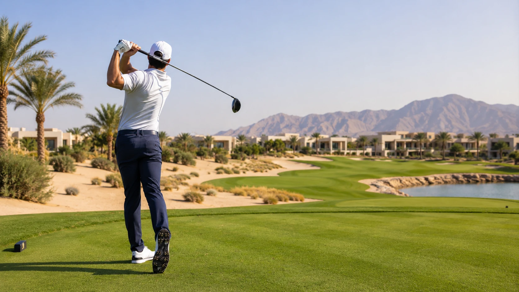 Male golfer in white shirt and navy trousers finishing a swing with a driver on a manicured green tee box, palm trees, sand, a lake, resort villas, and mountains behind under a clear blue sky.