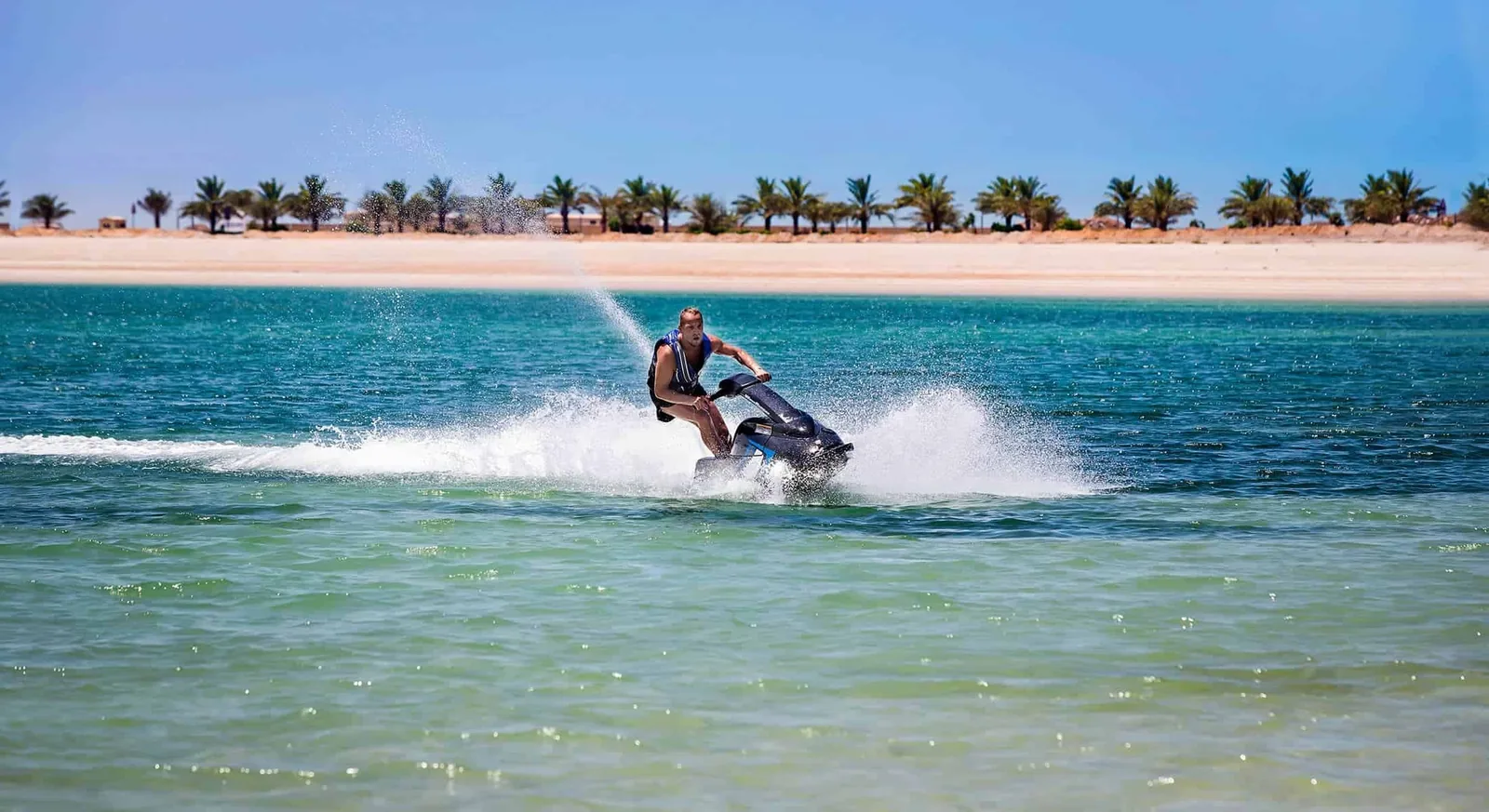 Man wearing a blue and black life vest rides a jet ski over turquoise water, spraying white foam, with a sandy beach and row of palm trees under a clear blue sky in the background.