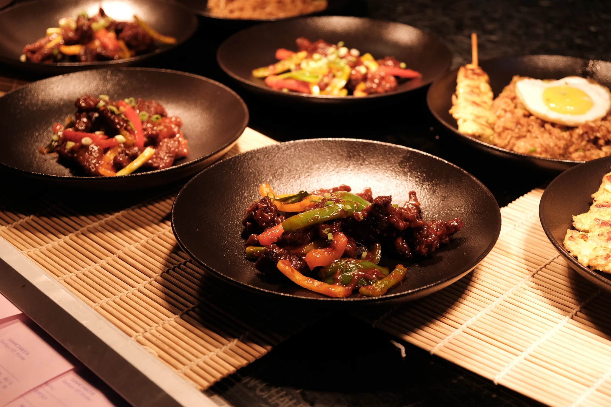 Sanchaya, Al Marjan Island, Ras Al Khaimah: Multiple black bowls filled with saucy stir-fried meat and strips of red, green, and orange bell pepper, arranged on a woven bamboo mat, with one bowl of fried rice topped with a fried egg and a grilled