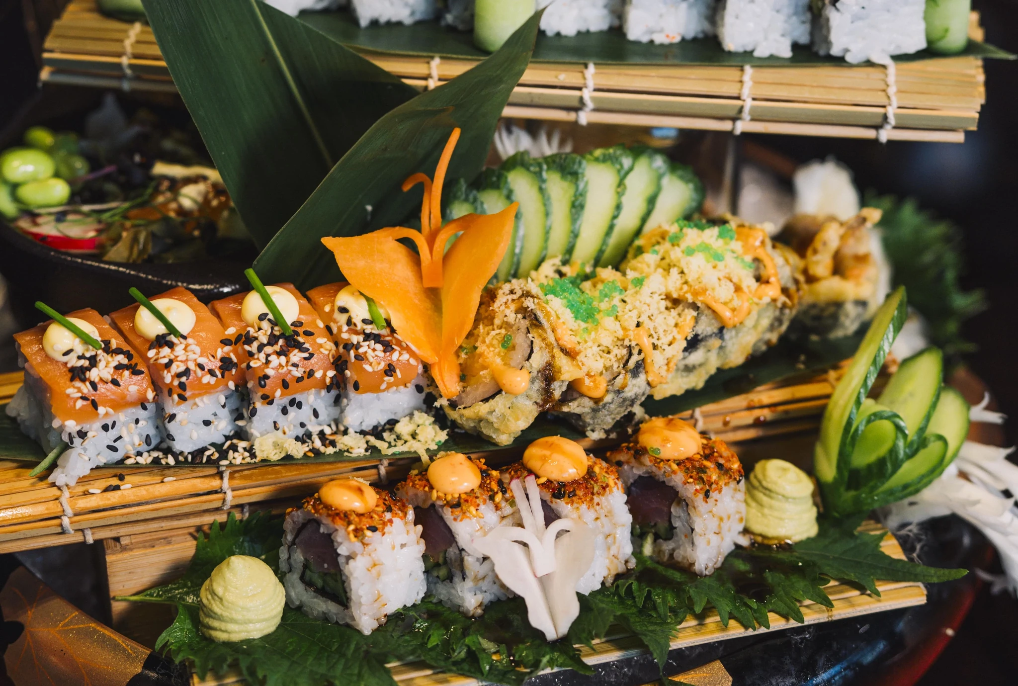 Multiple sushi rolls arranged on bamboo mats and green leaves, including salmon-topped rolls with sesame seeds, tempura crunch rolls with orange sauce and green roe, and tuna rolls with spicy mayo, garnished with sliced cucumber, a carved carrot flower, and wasabi mounds.