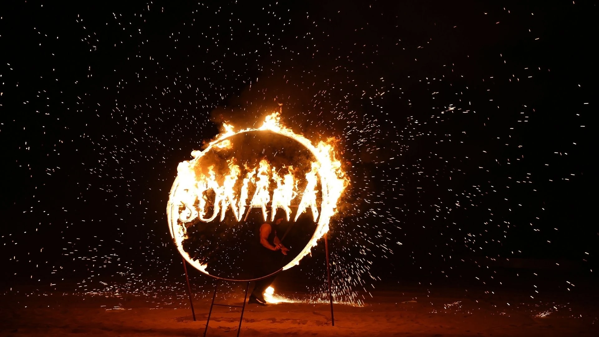 Sonara Camp Al Wadi, Ras Al Khaimah: Night fire performance featuring a large circular metal frame ablaze with flames spelling out letters, a silhouetted performer standing inside it, and sparks flying across a dark sandy beach.