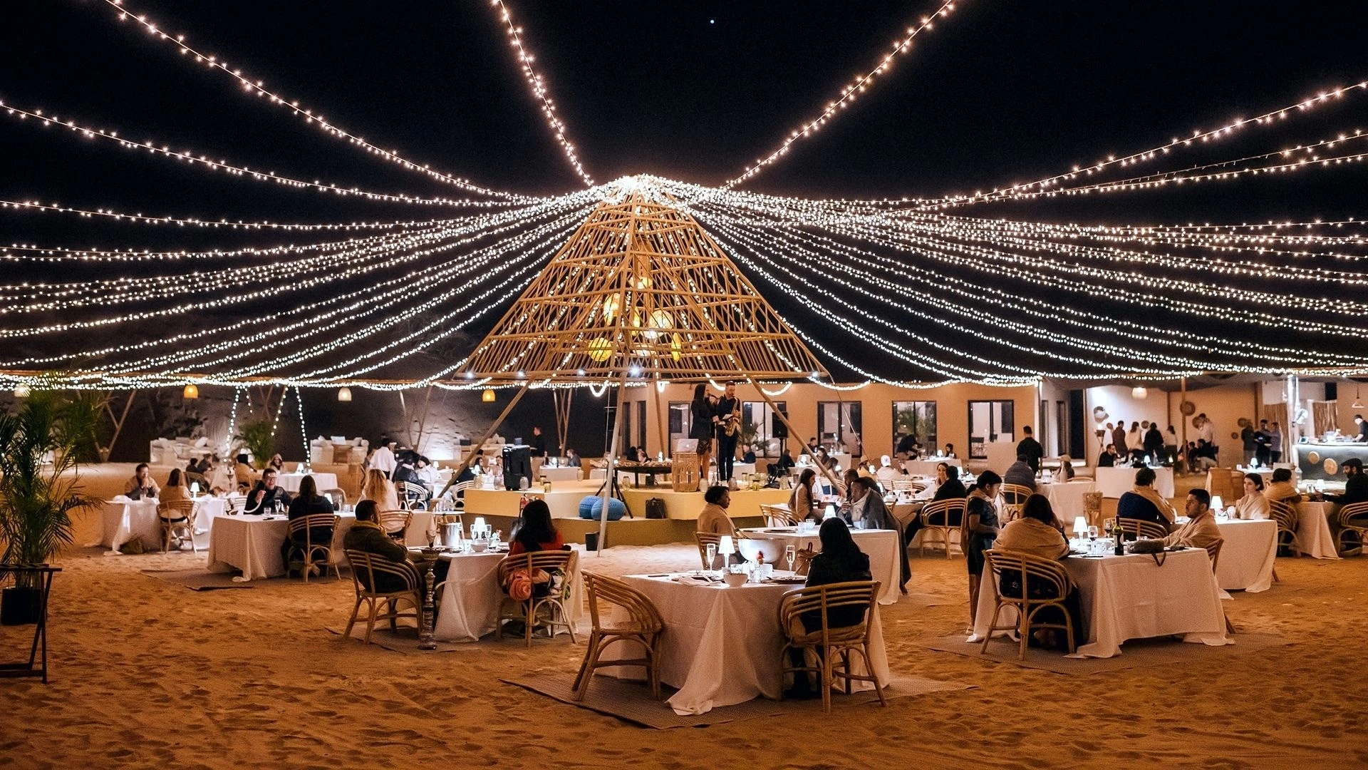 Sonara Camp Al Wadi, Ras Al Khaimah: Nighttime outdoor desert dining setup with dozens of guests seated at white-linen tables and rattan chairs on sand, illuminated by hundreds of warm string lights fanning out from a central bamboo pyramid frame,