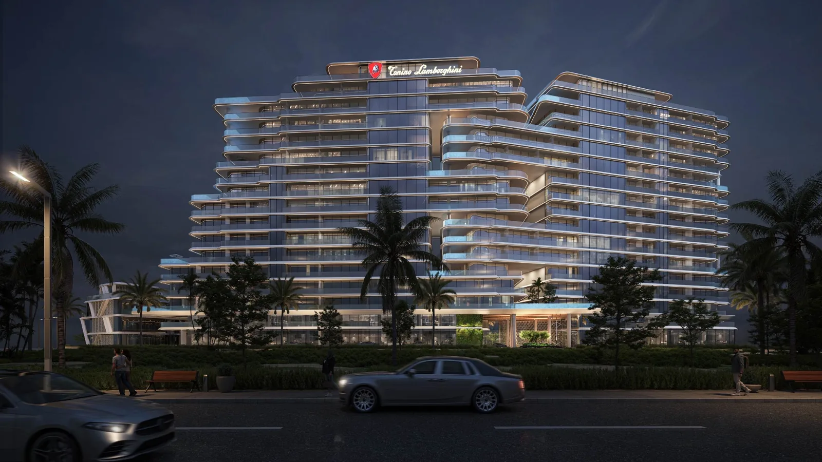 Nighttime view of a curved multi-story glass residential building with illuminated balconies and an illuminated Tonino Lamborghini sign on top, surrounded by palm trees, with cars driving along the road in the foreground and pedestrians on the sidewalk.