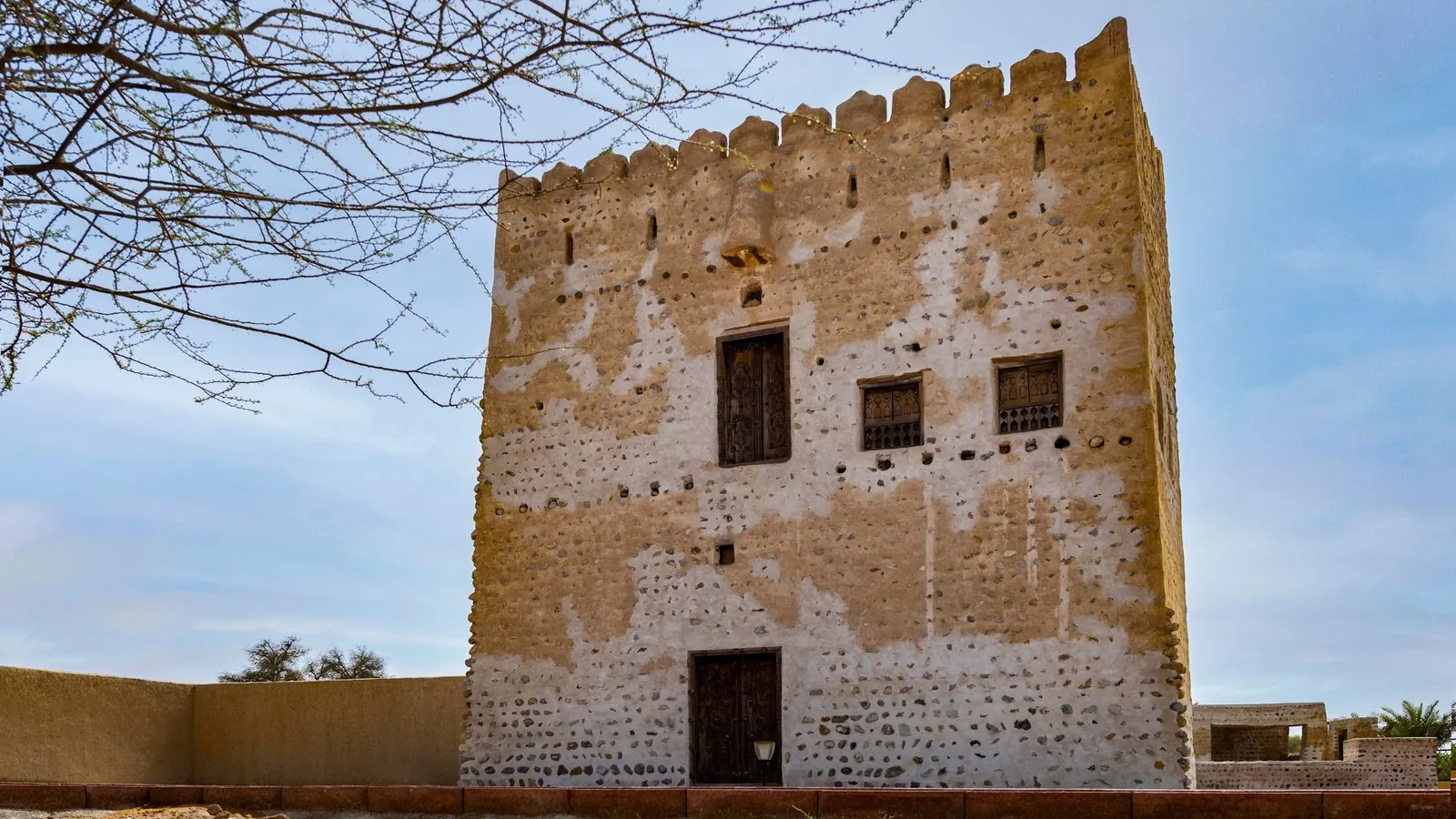 Old square fort tower built from stone and mud with a battlement top, carved wooden door and shuttered windows, framed by a bare tree branch and a low perimeter wall under a pale blue sky.