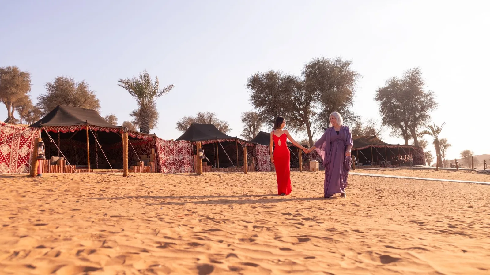 One woman in a red gown and another in a purple robe hold hands while walking on desert sand in front of black Bedouin tents draped with red patterned fabrics, framed by palm trees and desert shrubs under a bright sky.