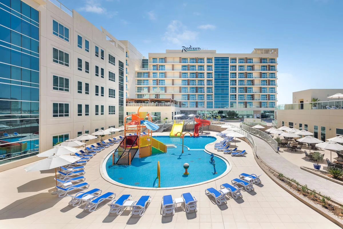 Radisson Resort Ras Al Khaimah Marjan Island, Al Hamra, Ras Al Khaimah: Outdoor hotel pool with a children's splash play structure and yellow and red water slides, surrounded by blue and white sun loungers and white parasols, with a multi-story