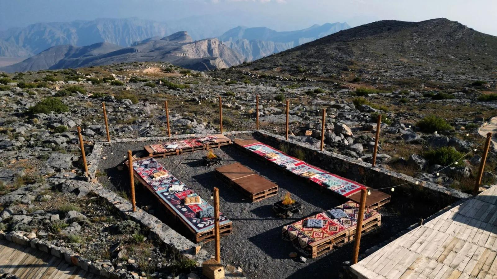 Outdoor seating area on a stony mountaintop with wooden pallet benches topped by red, blue, and patterned cushions arranged in a U shape around low tables and small fire pits, framed by timber posts with string lights and layered mountain peaks in the background.