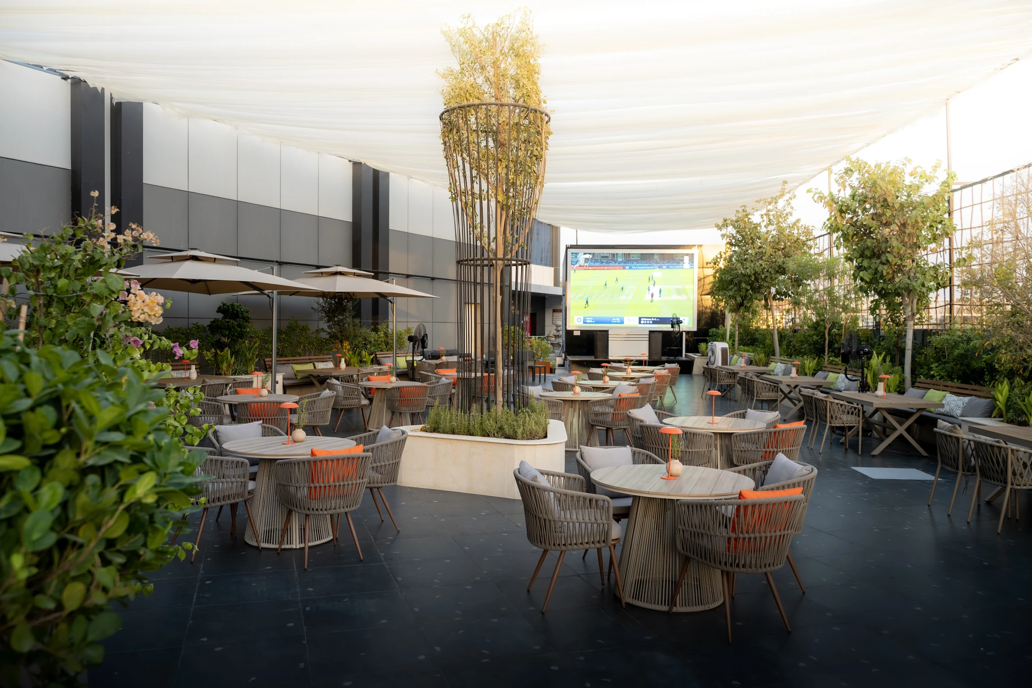 Outdoor terrace dining area with gray woven rope armchairs, round wooden tables topped with small orange lamps, potted plants and trees, tan patio umbrellas, and a large screen broadcasting a cricket match under a draped white canopy.