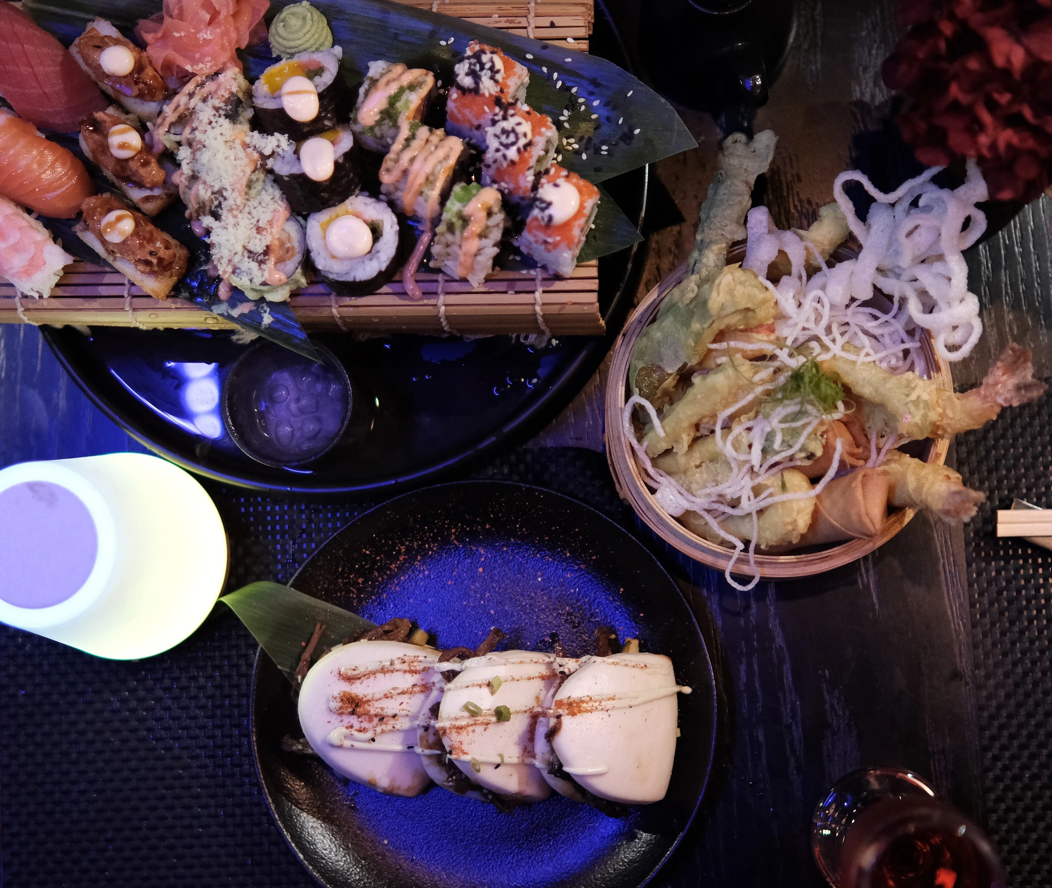 Sanchaya, Al Marjan Island, Ras Al Khaimah: Overhead view of a Japanese meal on a dark wooden table, including assorted sushi rolls and nigiri on a bamboo mat, a bamboo basket of tempura and spring rolls, three steamed bao buns drizzled with sauce