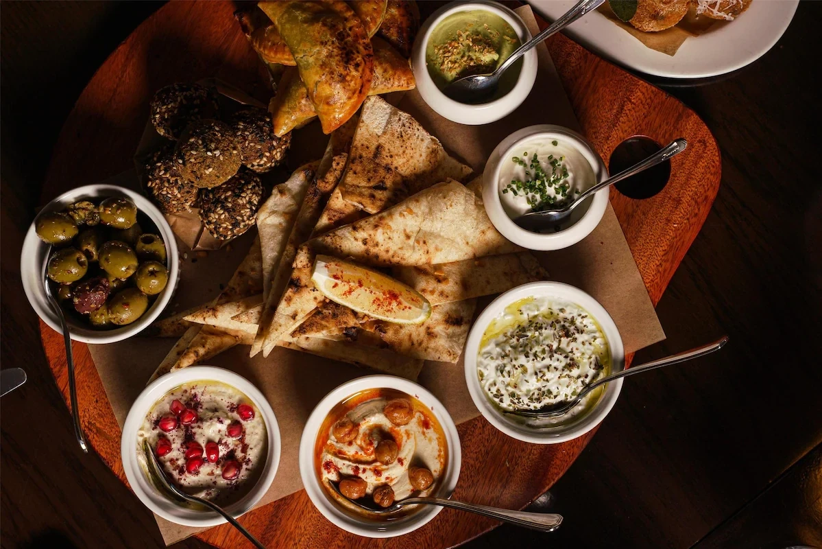 Overhead view of a mezze spread on a round wooden board, with several white bowls of dips including one topped with pomegranate seeds, one with chickpeas in chili oil, herbed yogurt, and green olives, surrounded by triangles of grilled flatbread, seeded crackers, and pastries.