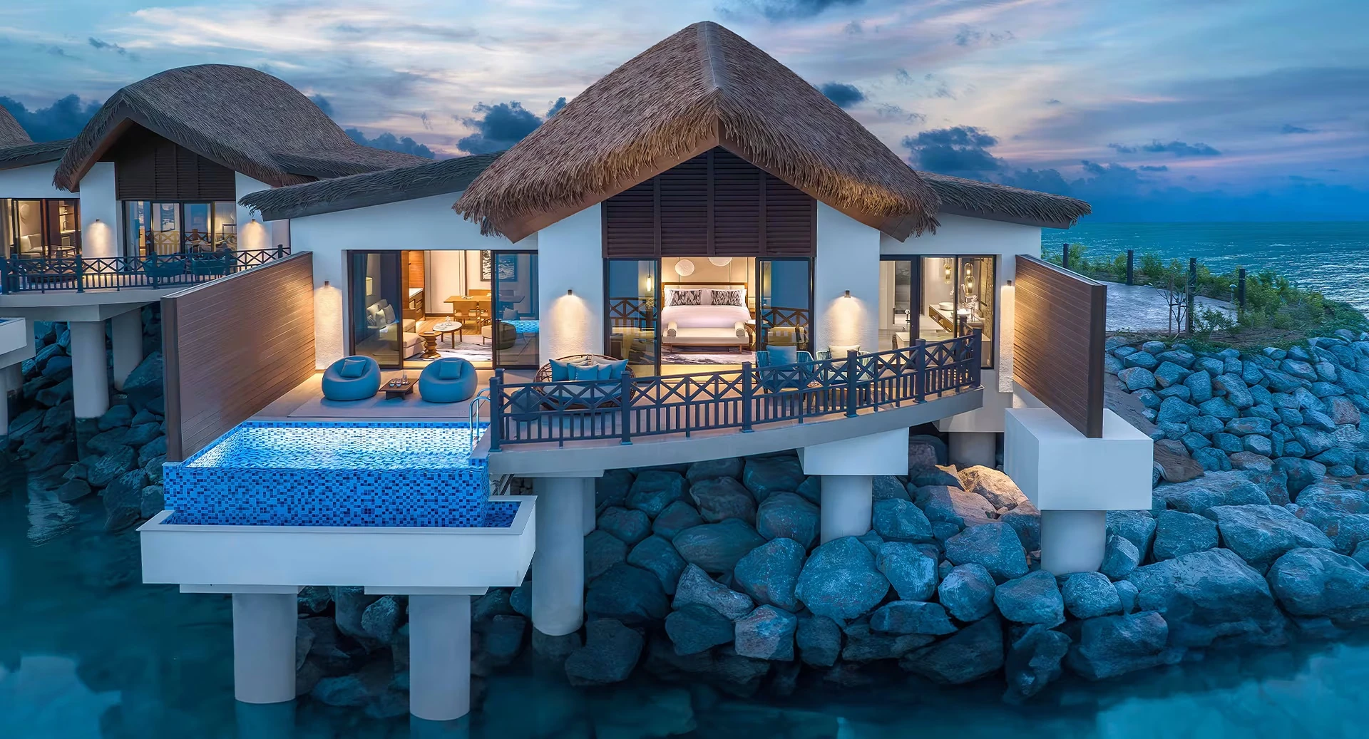 Anantara Mina Ras Al Khaimah Resort, Mina Al Arab, Ras Al Khaimah: Overwater villa with thatched roof, illuminated bedroom and living room, wooden deck with blue bean bag chairs, a private mosaic-tiled plunge pool, and rocks beside the sea at