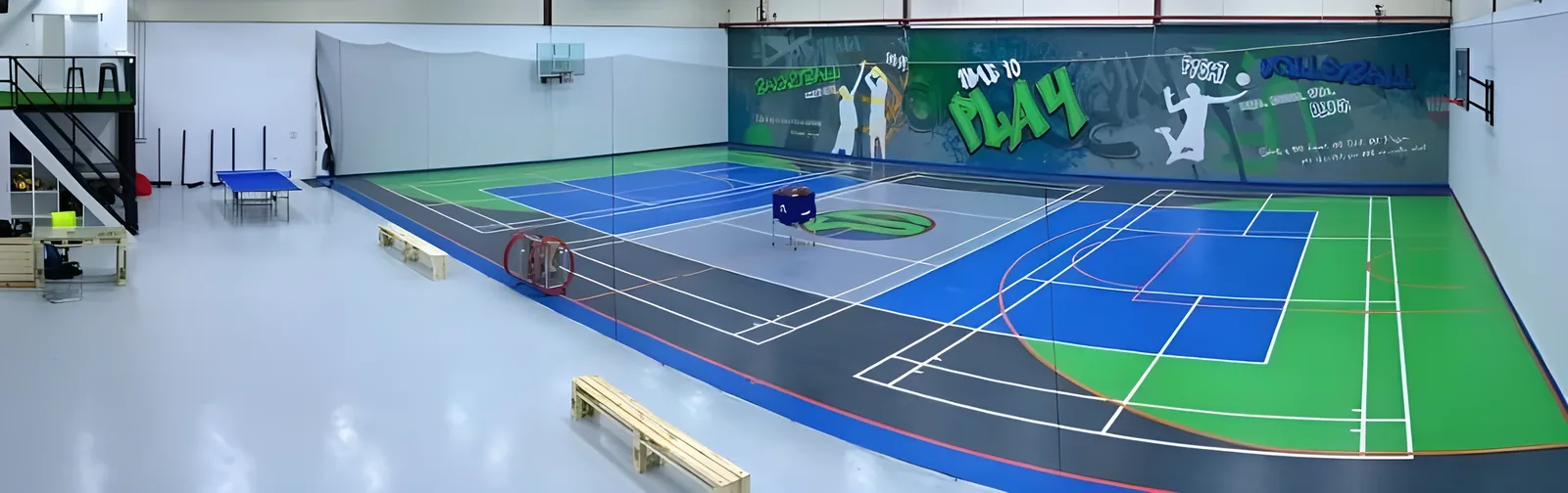 Panoramic view of an indoor sports facility featuring blue, gray, and green multi-purpose courts marked for badminton and basketball, with wooden pallets and a table tennis table along the sides and painted wall murals reading 'Time to Play' and 'Volleyball'.